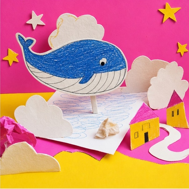 Paper-craft blue whale floating between clouds and stars above a tiny village